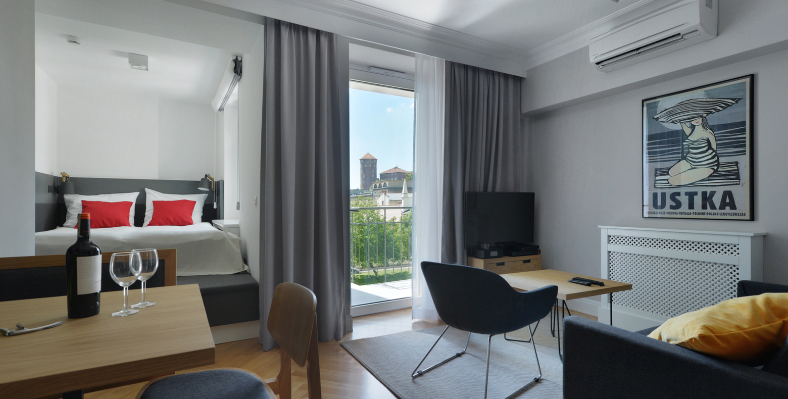 Studio hotel suite with alcove bed with red pillows, table with wine, lounge area, TV and balcony overlooking historic towers.