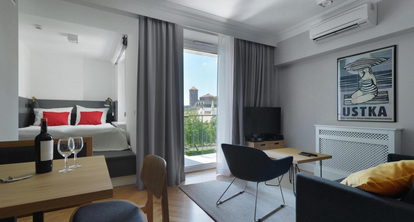 Modern studio with bed alcove and red pillows, dining table with wine, seating area and balcony overlooking historic towers.