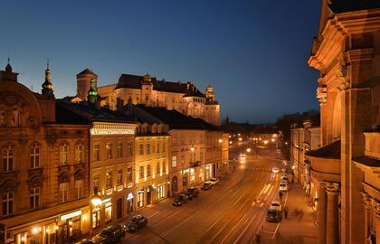 Historic hilltop castle and illuminated ornate facades above a lively evening street with boutique hotels, restaurants and tram tracks.