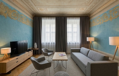 Elegant suite lounge with ornate painted ceiling, blue walls with gold frieze, large windows, gray sofa, armchairs and wooden coffee table.