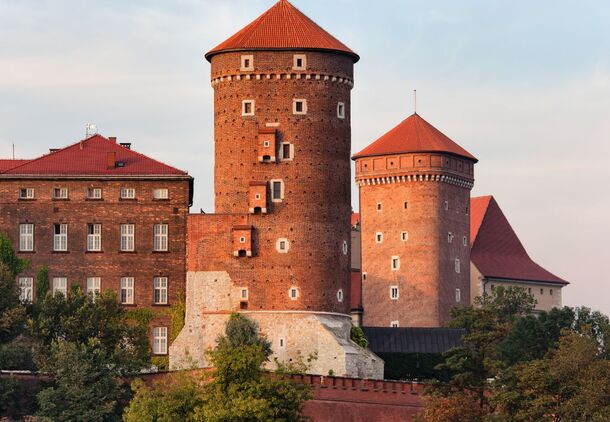 Historic red-brick castle towers with conical roofs above tree-lined ramparts at golden hour