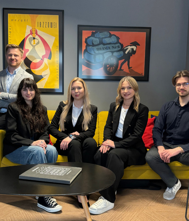 Pięcioro osób siedzi na żółtej sofie w nowoczesnym hotelowym salonie; plakaty na ścianie, stolik z książką, przyjazna, profesjonalna atmosfera