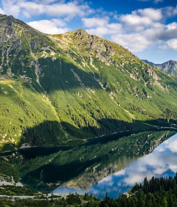 Górskie jezioro z odbiciem szczytów, zalesionym brzegiem i schroniskiem w dali — idealne na wędrówki i widoki.