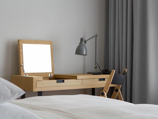 Wooden vanity desk with mirror, adjustable desk lamp and chair by a neatly made bed and grey curtains in a modern hotel room