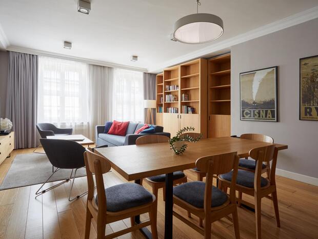 Bright modern suite living and dining area with wooden table and chairs, cozy sofa with red cushions, TV, bookshelf and large windows with curtains.