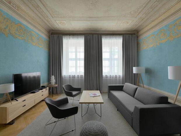 Spacious hotel sitting room in a suite with ornate ceiling, teal walls with gold frieze, modern gray sofa, armchairs, and TV.