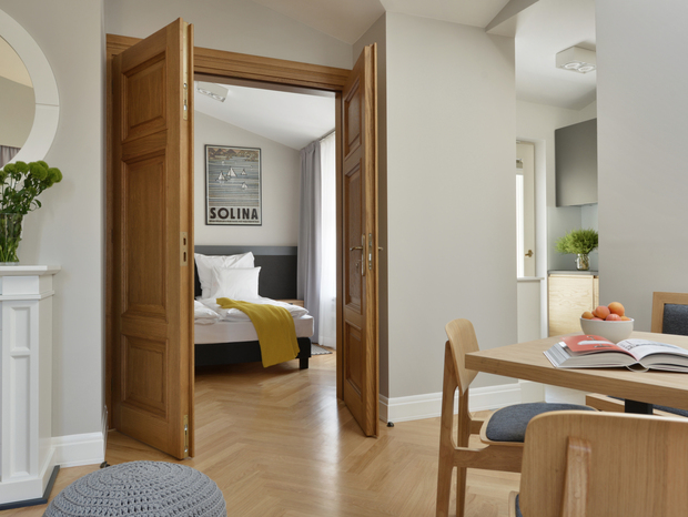 Bright modern suite with wooden double doors to a bedroom with white linens and yellow throw, dining table and fireplace.