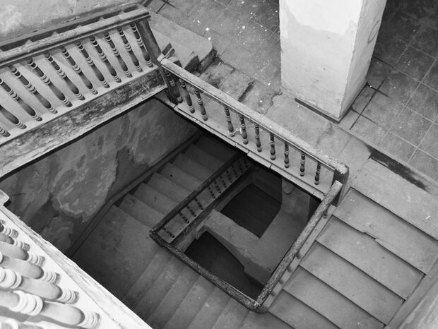 Zniszczona drewniana balustrada i kamienne schody w zabytkowym budynku, widok z góry na patynę i płytki podestu.