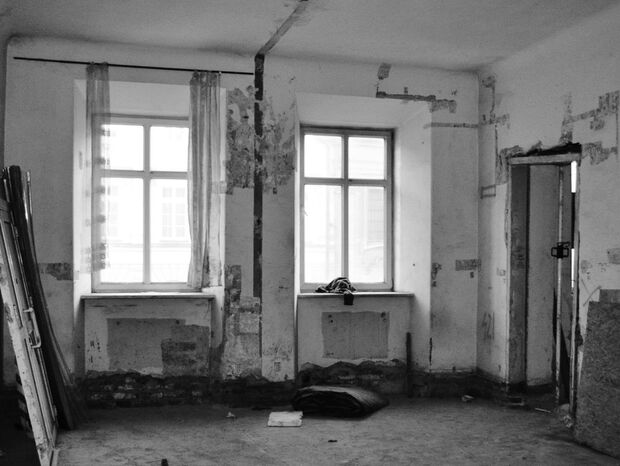 Puste pomieszczenie w remoncie z odpadającym tynkiem, dwoma oknami, drzwiami opartymi o ścianę, odsłoniętą cegłą i metalowym słupem