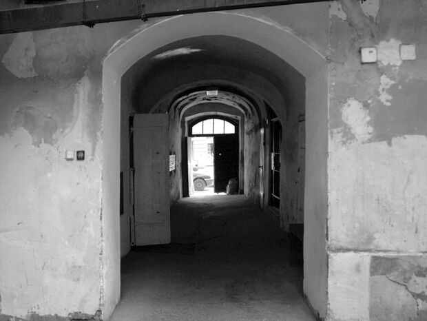 Łukowe przejście w starym budynku, z odpadającym tynkiem, ciemny korytarz otwiera się na jasną ulicę z zaparkowanym samochodem.