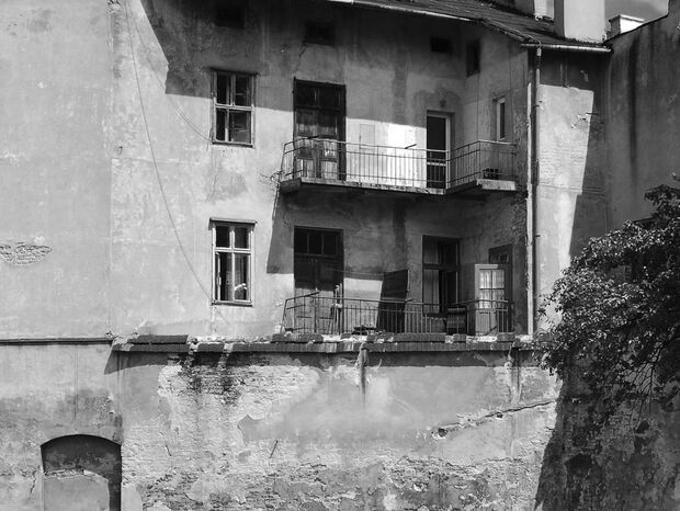 Zniszczona fasada kamienicy z dwoma balkonami, otwartymi drzwiami, odpadającym tynkiem, metalowymi balustradami i drzewem po prawej