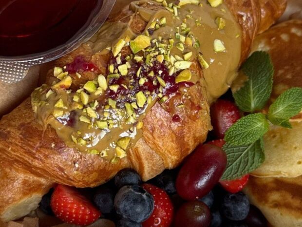 Maślany croissant z karmelem, konfiturą i posiekanymi pistacjami, podany z owocami, mini naleśnikami, miętą i syropem.