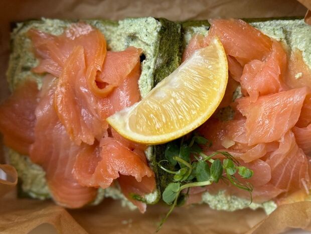 Wędzony łosoś na zielonym chlebie z ziołowym serkiem, ćwiartką cytryny i mikroliśćmi, podany w papierze.