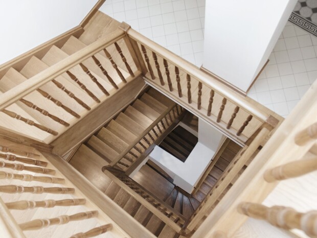 Jasne dębowe schody wewnętrzne z toczonymi balustradami schodzące przez kwadratowy szyb, widoczny jasny kafelkowany hol.