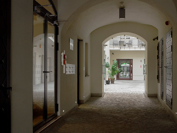 Sklepione przejście z brukowaną posadzką prowadzi do jasnego dziedzińca z donicami, szklanymi drzwiami i balkonem — spokojne wejście hotelu