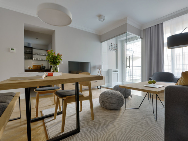 Jasny nowoczesny apartament z otwartą częścią dzienną i jadalnią, drewniane meble, sofa, pufy, telewizor i balkon.