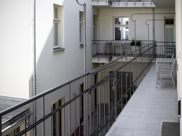 Korytarzowy balkon z kaflową posadzką, metalową balustradą i ławką, widok na spokojne wewnętrzne patio hotelu.