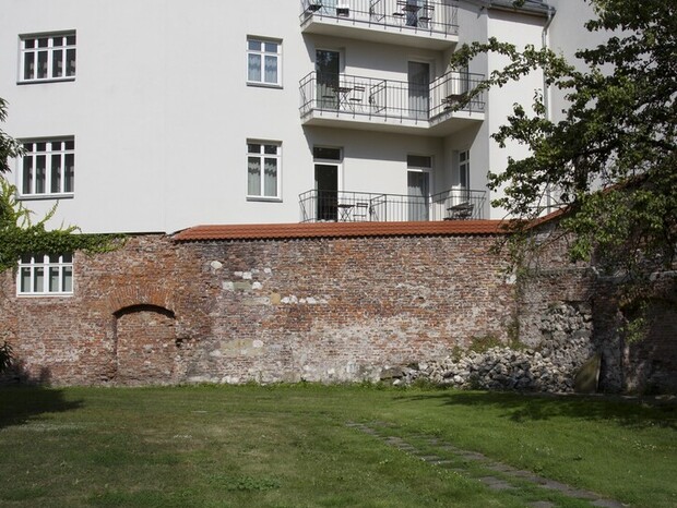 Spokojne podwórko z trawnikiem, zabytkowym murem z cegły i białym budynkiem z balkonami i stolikami.