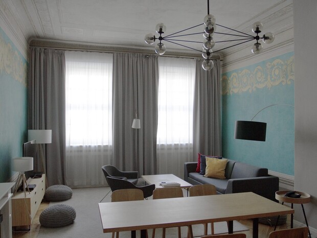 Apartamentowy salon z wysokim zdobionym sufitem, turkusowymi ścianami, nowoczesnym żyrandolem, sofą z kolorowymi poduszkami i stołem jadalnym