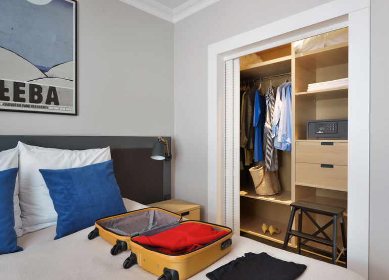 Bright modern hotel room: bed with blue pillows, open yellow suitcase on bed, open wardrobe with hanging clothes, in-room safe and folding stool.