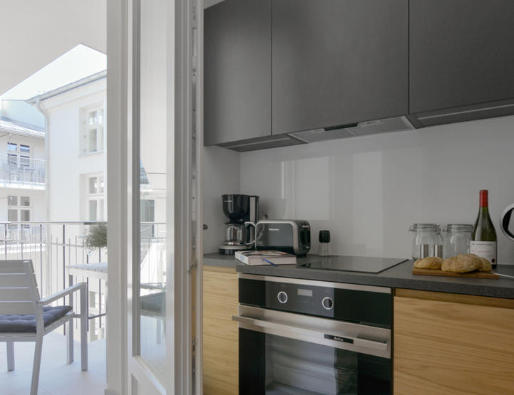 Modern kitchenette in a suite with wood cabinets, built-in oven and hob, coffee maker and toaster, bread and wine on counter, balcony seating and daylight.