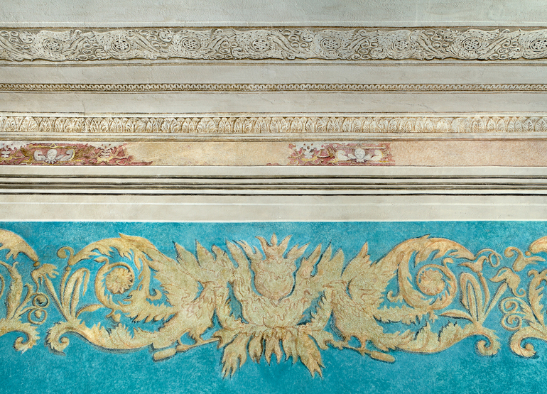 Ornate plaster cornice and carved molding above a turquoise wall frieze with gold leaf floral and scrolling motifs.