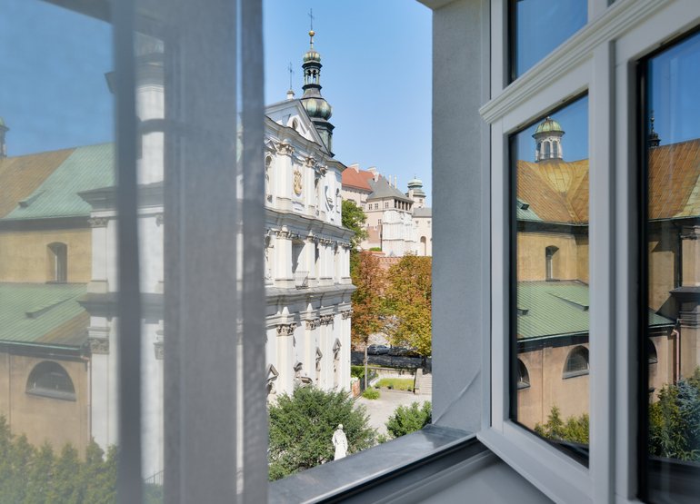 Open hotel window with sheer curtain framing historic baroque church, copper domes, tree-lined courtyard and statue below.