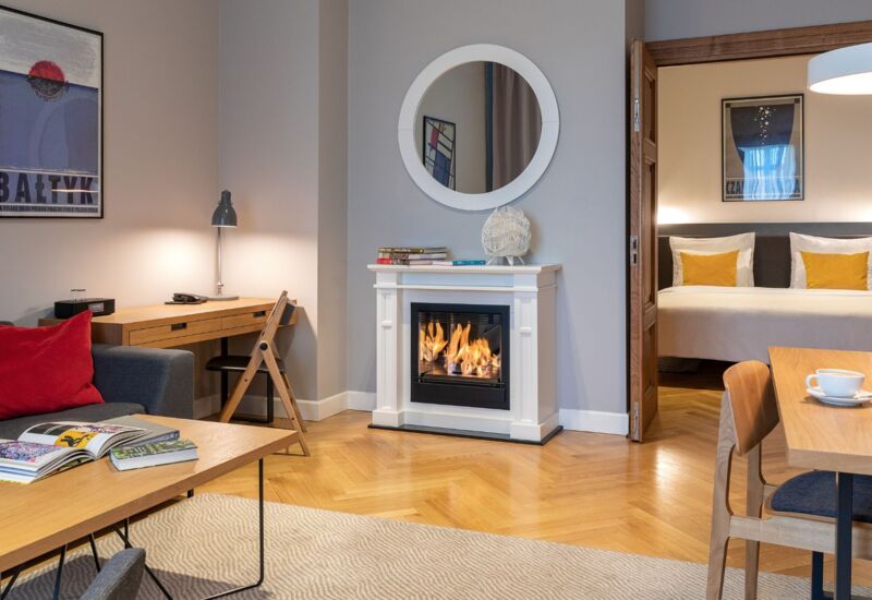 Przytulny apartament z palącym się kominkiem, częścią wypoczynkową, biurkiem i stołem jadalnym; przyległa sypialnia z łóżkiem dwuosobowym i żółtymi poduszkami.