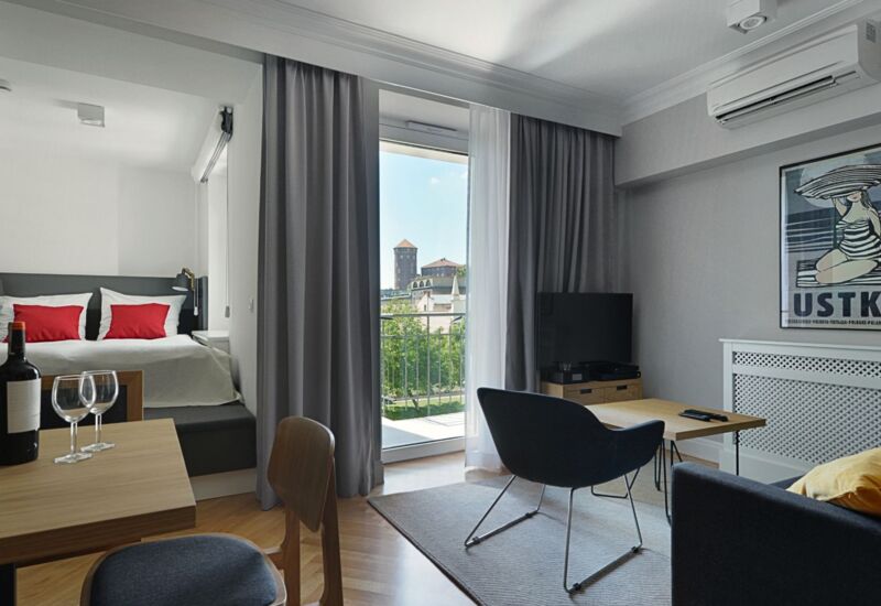 Jasne studio z podwójnym łóżkiem i czerwonymi poduszkami, balkon z widokiem na wieżę, część wypoczynkowa, TV, wino.