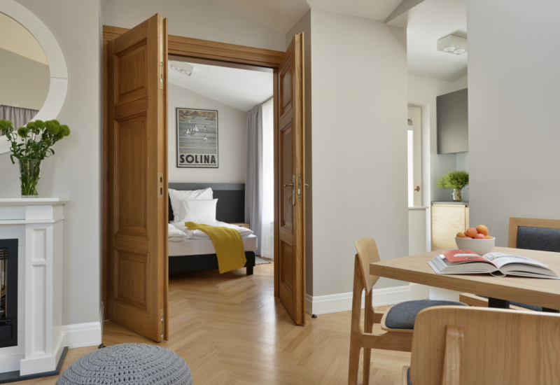 Jasny, nowoczesny apartament z drewnianymi drzwiami do sypialni z białą pościelą i żółtym pledem, stołem jadalnym i kominkiem