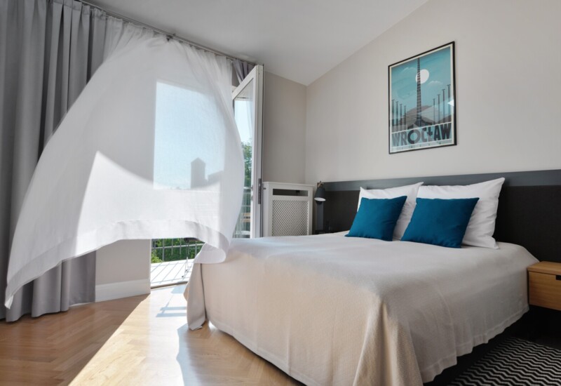 Jasny, nowoczesny pokój dwuosobowy z balkonem i falującą lekką zasłoną, łóżko z niebieskimi poduszkami, lampka nocna i drewniana podłoga