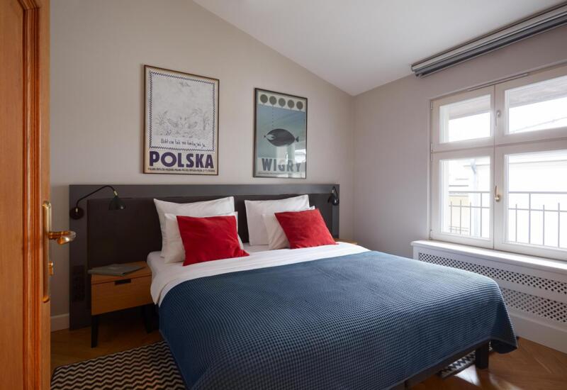 Jasny pokój gościnny z podwójnym łóżkiem, granatową narzutą i czerwonymi poduszkami, lampkami nocnymi, plakatami i dużym oknem