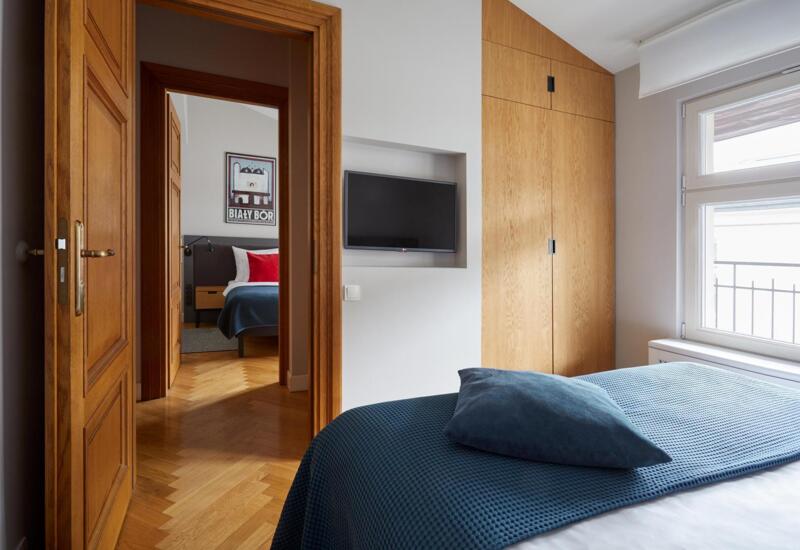 Połączone pokoje gościnne z ciepłymi drewnianymi drzwiami, parkietem, telewizorem w niszy, szafą, niebieską narzutą i oknem na balkon.