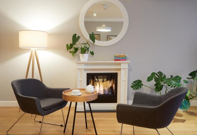 Przytulny kącik z dwoma nowoczesnymi fotelami, stolikiem z imbrykiem, palącym się kominkiem, lampą i roślinami.
