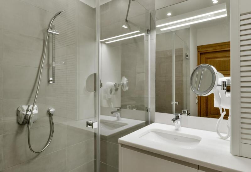 Nowoczesna łazienka hotelowa z przeszkloną kabiną prysznicową, chromowaną słuchawką, białą umywalką, podświetlanym lustrem i suszarką ścienną