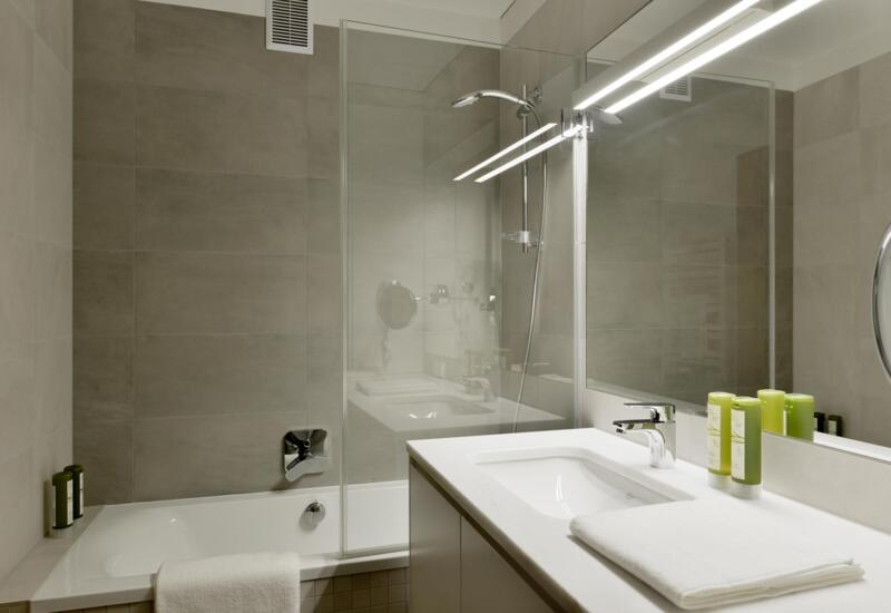 Nowoczesna łazienka hotelowa z wanną i szklaną ścianką prysznicową, podświetlanym lustrem, białą umywalką, chromowanymi bateriami, ręcznikiem i zielonymi kosmetykami