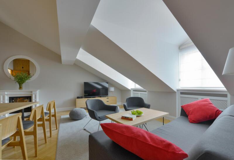 Salon w apartamencie na poddaszu z pochylonymi oknami, szarą sofą i czerwonymi poduszkami, fotelami, stołem, TV i kominkiem.