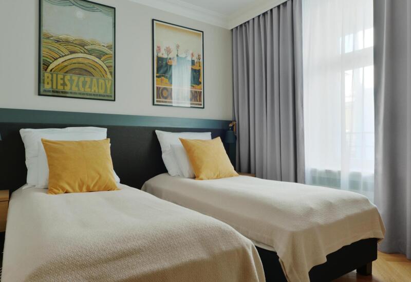 Pokój z dwoma pojedynczymi łóżkami, musztardowymi poduszkami, jasną pościelą, plakatami, lampkami nocnymi i światłem przez cienkie zasłony.