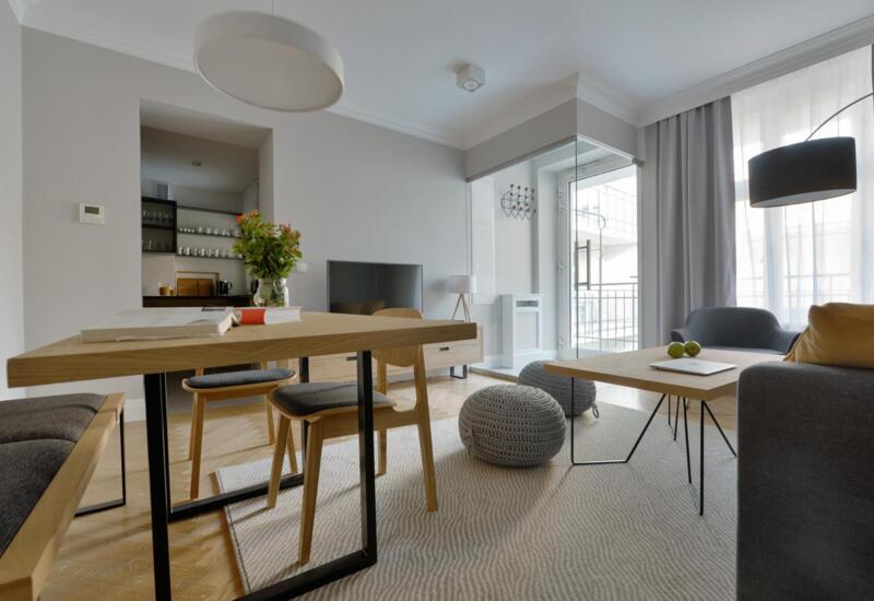 Nowoczesny apartament: jadalnia z dębowym stołem, wygodna sofa, plecione pufy, telewizor i balkon z naturalnym światłem.
