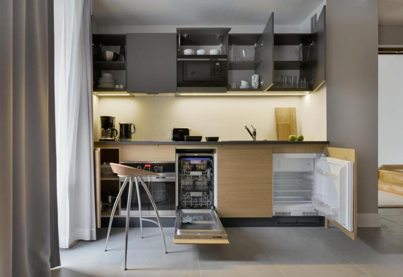 Nowoczesny aneks kuchenny w apartamencie z mikrofalówką, ekspresem do kawy, czajnikiem, zlewem, otwartą zmywarką i lodówką, drewnianymi szafkami i stołkiem barowym.