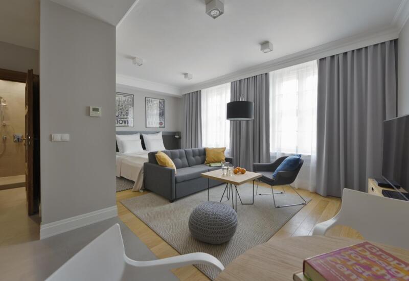 Nowoczesne studio z podwójnym łóżkiem, szarą sofą i fotelem, stolikiem z napojami i pomarańczami oraz dużymi oknami i szarymi zasłonami