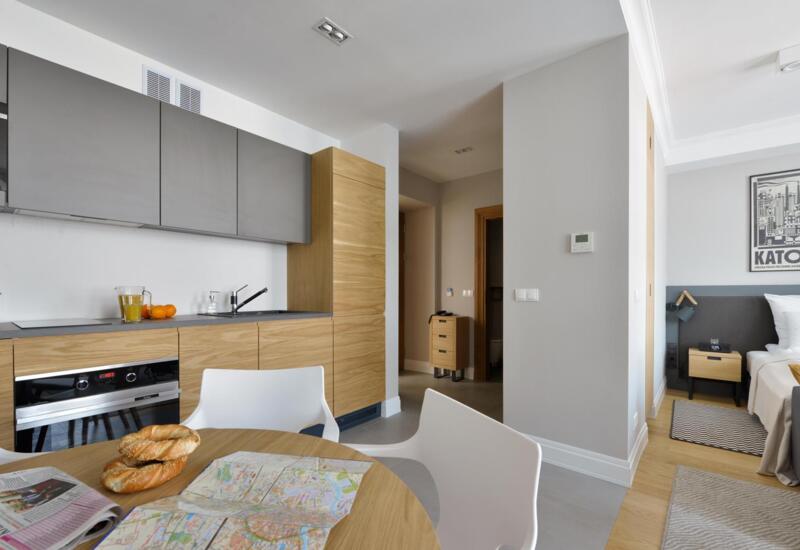 Nowoczesne studio z aneksem kuchennym, drewnianymi szafkami, stołem z mapą i wypiekami, łóżko i stolik nocny.