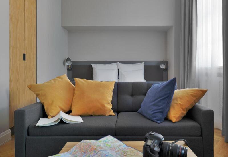 Nowoczesne studio z szarą sofą i musztardowo‑niebieskimi poduszkami, łóżkiem z białymi poduszkami, drewnianą szafą, mapą i aparatem na stoliku