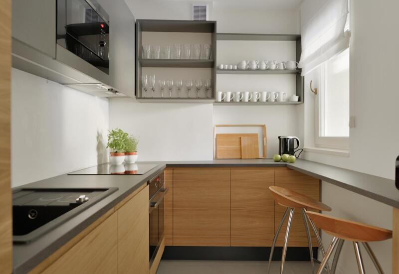 Nowoczesny aneks kuchenny z drewnianymi szafkami i ciemnym blatem, płytą i piekarnikiem, barem z dwoma stołkami, otwarte półki, czajnik, zioła