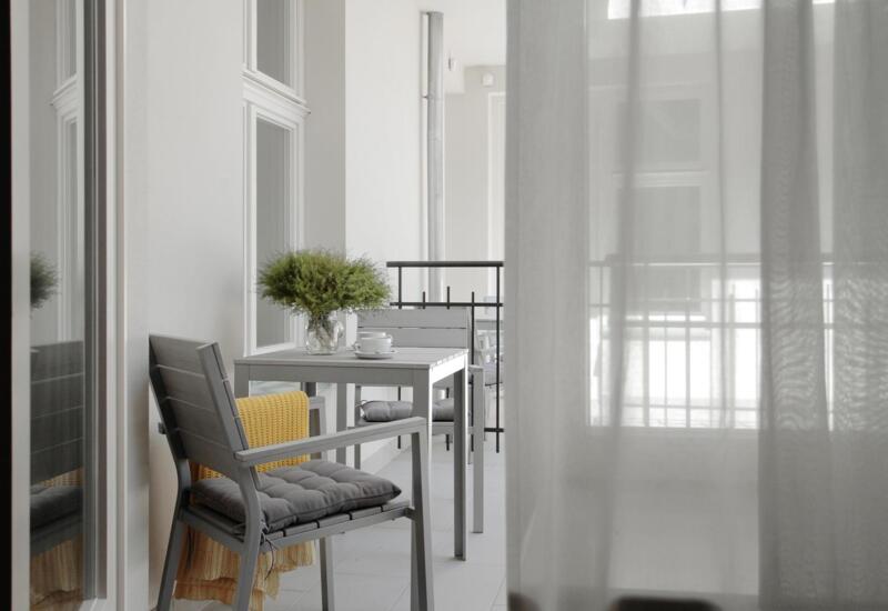 Przytulny balkon hotelowy z szarym stolikiem, poduszkami na krzesłach, żółtym kocem, zielonym bukietem i lekkimi zasłonami.