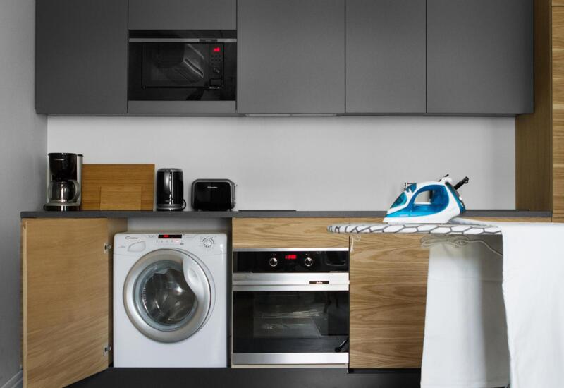Kompaktowy aneks z pralką, piekarnikiem, mikrofalą, ekspresem do kawy, tosterem, czajnikiem i żelazkiem na desce.