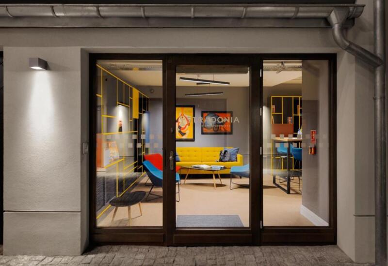 Nowoczesny hotelowy salon widoczny przez szklane drzwi; żółta sofa, kolorowe fotele, bar z hokerami i przytulne oświetlenie