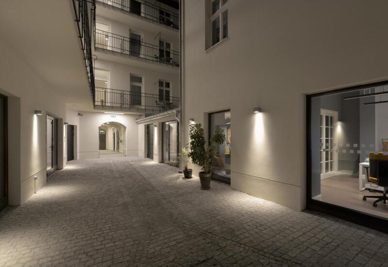 Spokojne, oświetlone podwórze hotelowe z brukiem, kinkietami, donicami i balkonami; przez szybę widać parterową strefę wypoczynkową.