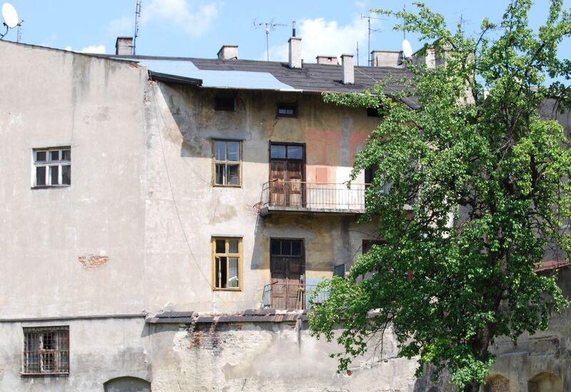 Zniszczona elewacja podwórza z odchodzącym tynkiem, dwiema małymi balkonami z żelaznymi balustradami, drewnianymi oknami i dużym zielonym drzewem.