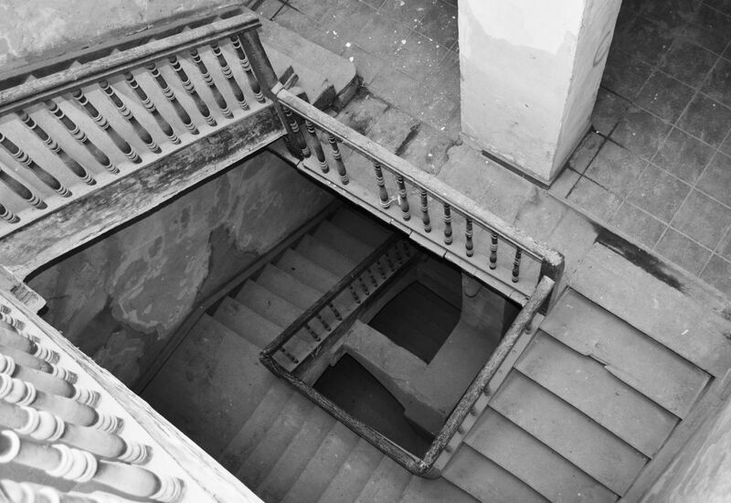 Zniszczona drewniana balustrada i kamienne schody w zabytkowym budynku, widok z góry na patynę i płytki podestu.